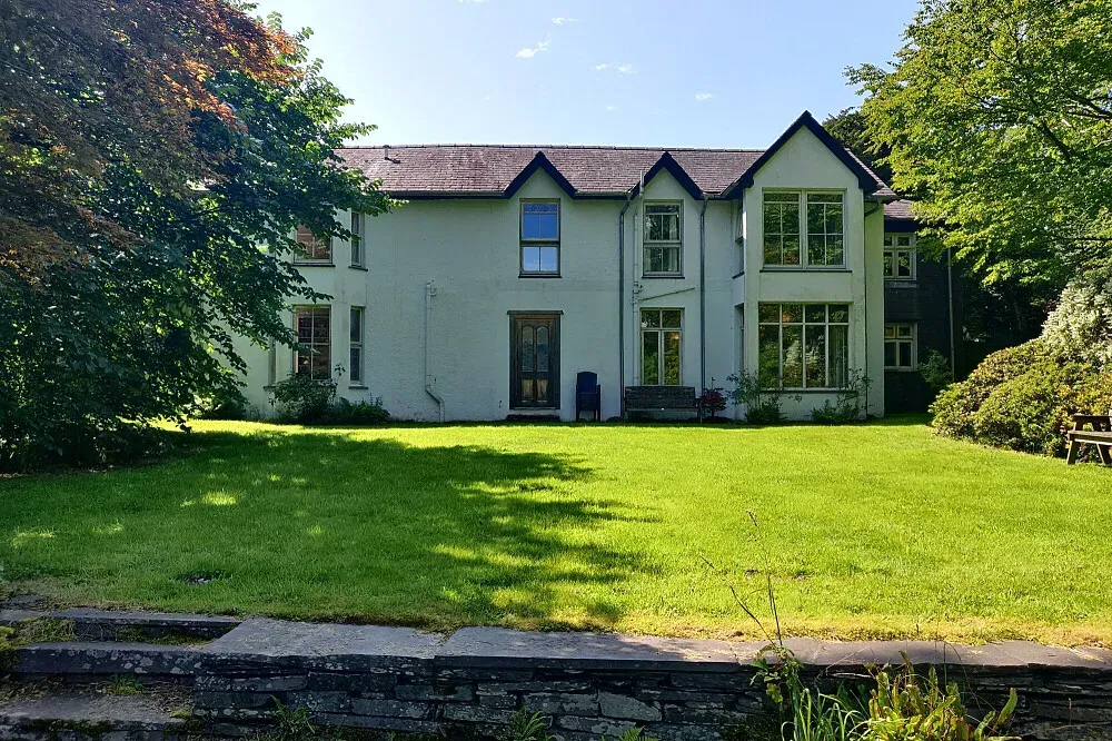 Trigonos Retreat Centre — The center is situated within 18 acres of varied natural beauty, featuring meticulously kept gardens, expansive meadows, and small groves of trees. A stream cascades gently through the property, eventually feeding into a crystal-clear lake, all set against the dramatic backdrop of the Snowdonia mountains. This creates a deeply serene and visually inspiring atmosphere.